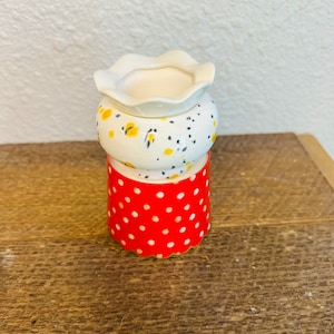 May include: A white ceramic planter with a red and white polka dot base. The planter has a scalloped rim and a floral pattern with yellow and black accents.