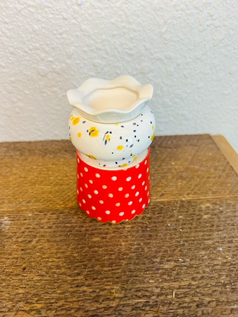 May include: A white ceramic planter with a red and white polka dot base. The planter has a scalloped rim and a floral pattern with yellow and black accents.