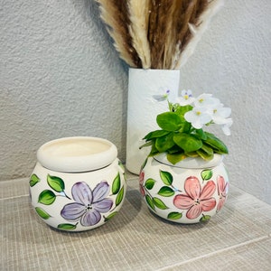 May include: Two small, round, white ceramic pots with hand-painted floral designs. One pot features purple flowers, the other pink. Green leaves and stems accent the flowers. The pots sit on a light-colored wooden surface.