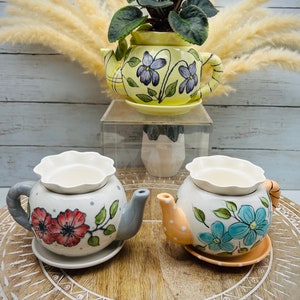 May include: Three ceramic teapot planters with floral designs. The top planter is yellow with purple flowers and a green plant. The bottom two planters are white with pink and blue flowers. The planters are on a wooden surface.