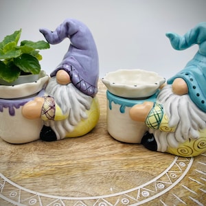 May include: Two ceramic gnome planters with yellow and white paint details. One planter has a purple hat and holds a small green plant. The other planter has a blue hat and is empty.