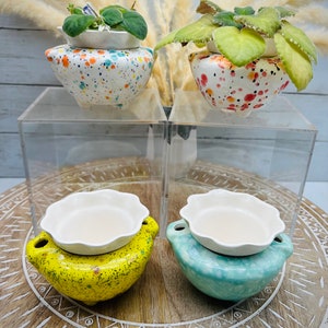 May include: Four ceramic planters with different colored glazes. Two planters are white with colorful speckles, one is yellow with green speckles, and one is blue with white speckles. The planters are on a wooden surface with a decorative pattern.