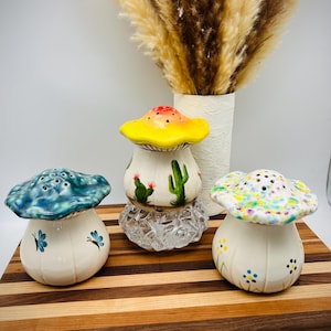 May include: Three ceramic mushroom-shaped salt and pepper shakers. One has a yellow cap with red spots, another has a blue cap, and the third has a floral design. The shakers are on a wooden cutting board with a vase of dried flowers in the background.