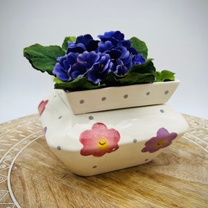 May include: A white ceramic planter with a square shape and a floral design. The planter has a smaller, white, polka-dotted planter on top. The planter is filled with a purple African violet plant.