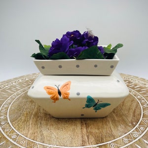 May include: A two-piece cream-colored ceramic container with a lid, decorated with orange and teal butterflies and gray polka dots. The top container holds purple flowers and green leaves. The set is placed on a wooden surface.