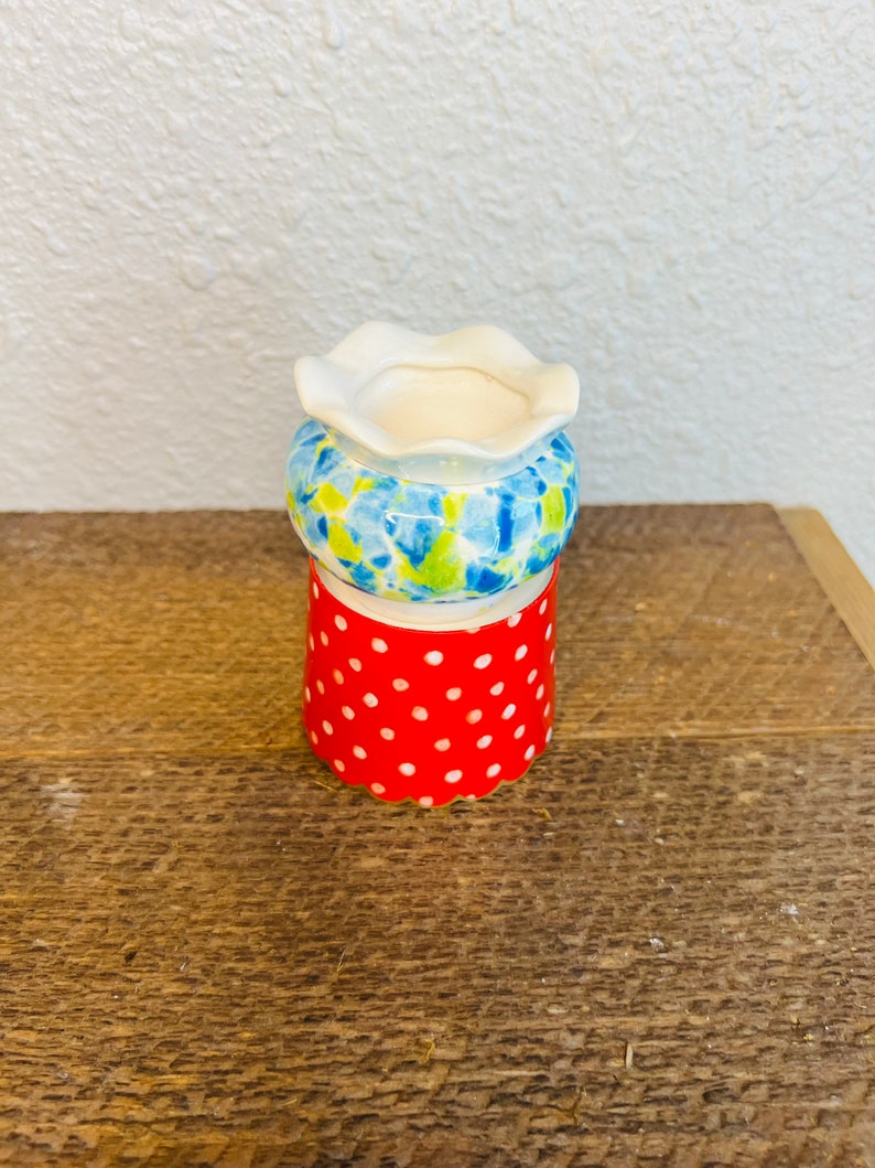 May include: A small white ceramic vase with a scalloped rim. The vase is decorated with a blue and green floral pattern and a red polka dot pattern.