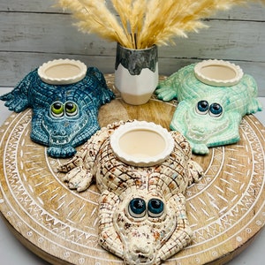 May include: Three ceramic alligator candle holders on a wooden tray with a white, brown, and blue design. The alligator candle holders have a white, blue, and green glaze. The alligator candle holders have a round, white candle dish on top.