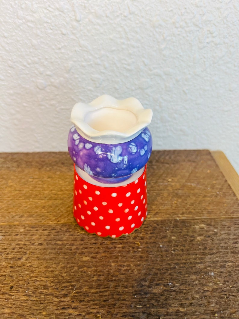 May include: A small purple and white ceramic planter with a white rim sits on a red and white polka dot base.
