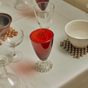Puede incluir: Un mantel blanco con una variedad de cristalería, incluyendo una copa de vino tinto, una copa de vino transparente y una copa con tallo transparente. También hay un tazón pequeño con cuentas marrones y un tazón blanco sobre un posavasos de cuentas plateadas.