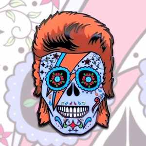 Puede incluir: Un pin de esmalte colorido que presenta una calavera de azúcar con un diseño de rayo, que recuerda el look icónico de David Bowie. La calavera está decorada con intrincados patrones florales y colores brillantes, incluyendo rojo, azul, verde y naranja.