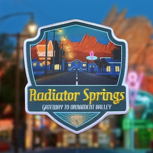 Può includere: Toppa ricamata raffigurante Radiator Springs di notte, con la scritta "Radiator Springs" in giallo. La toppa ha la forma di uno scudo con un bordo bianco e include la scritta "Gateway to Ornament Valley".