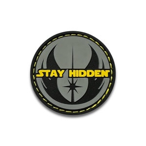 May include: A black and gray round patch with a yellow stitched border. The patch features a stylized symbol with a star in the center and the text "STAY HIDDEN" in yellow letters.