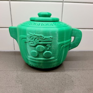 May include: A green ceramic teapot with a dragon design and a lid. The teapot has a rounded body and a spout on one side. The dragon design is embossed on the front of the teapot.