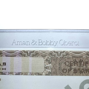 May include: A close-up of a document with the text "Aman & Bobby Oberoi" printed in silver lettering. The document has a decorative pattern and a blank space for a signature.