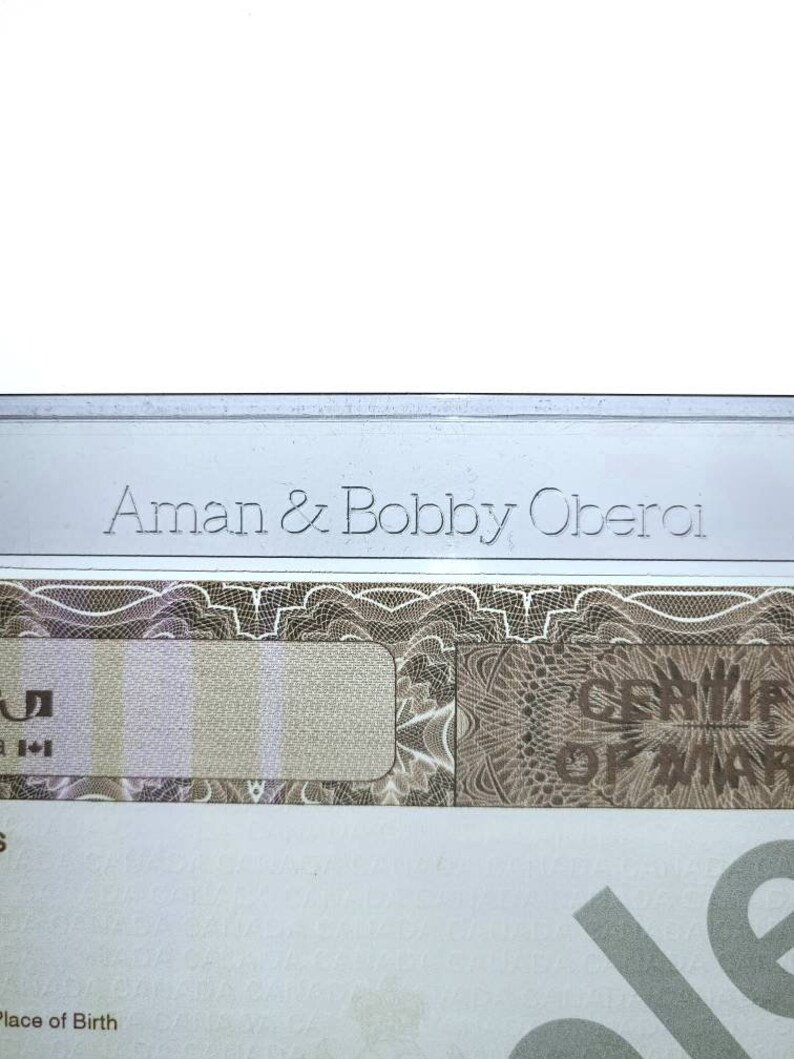 May include: A close-up of a document with the text "Aman & Bobby Oberoi" printed in silver lettering. The document has a decorative pattern and a blank space for a signature.