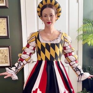 Könnte beinhalten: Ein historisch anmutendes Kleid mit einem Oberteil im gelb-schwarzen Rautenmuster, langen Ärmeln mit weißen und roten Streifenakzenten sowie einem weiten Rock mit vertikalen roten, weißen und schwarzen Bahnen. Dazu wird eine passende Kopfbedeckung getragen.