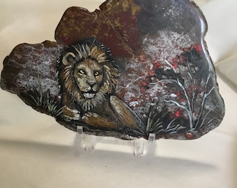 May include: A hand-painted stone sculpture of a lion with a brown and black mane, lying down on a rock with a background of red and white flowers.