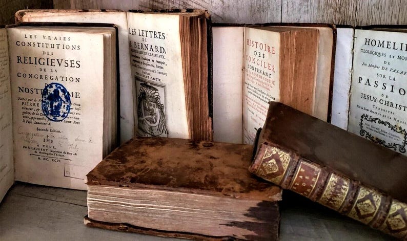 May include: A collection of antique books with aged, textured covers. The spines and pages show signs of wear, with titles in French. One book features a blue seal. The books are arranged on a wooden surface, evoking a sense of history and scholarly interest.