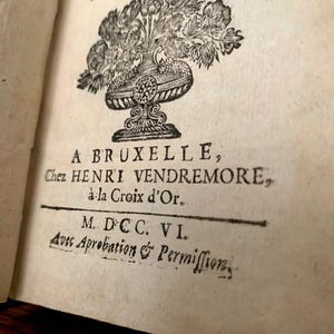 May include: An open book page with antique text and illustrations. The page features a decorative floral illustration above the text "A BRUXELLE". Additional text includes "Chez HENRI VENDREMORE" and "M. DCC. VI."