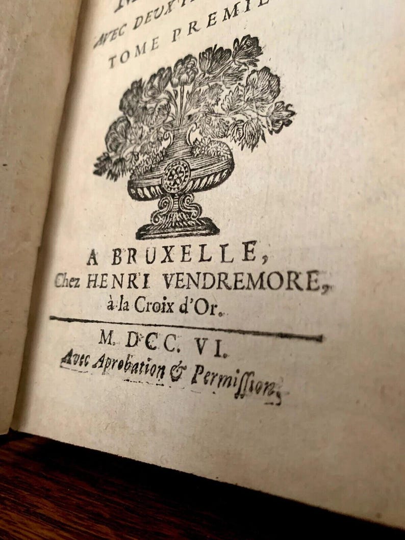 May include: An open book page with antique text and illustrations. The page features a decorative floral illustration above the text "A BRUXELLE". Additional text includes "Chez HENRI VENDREMORE" and "M. DCC. VI."