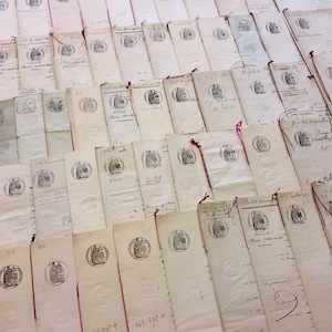 May include: A collection of aged, cream-coloured documents, many with a circular seal featuring a ship. The documents are arranged in a layered pattern, some with red string ties. Handwritten text and markings are visible.
