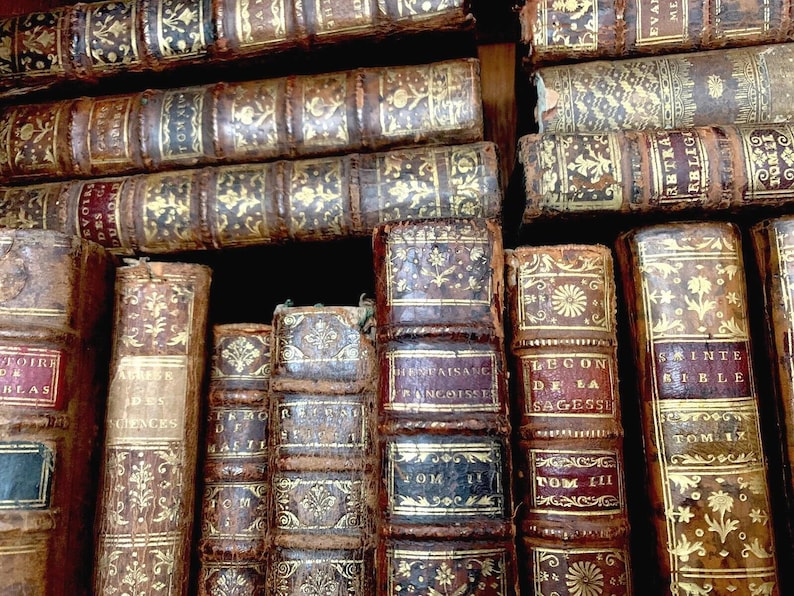May include: A selection of antique books with worn leather covers. The spines and covers display gold embossed patterns and titles. The books are stacked, revealing their historical and decorative appeal. The aged appearance suggests a rich history.