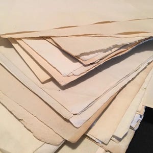 以下が含まれることがあります： ざらざらした、破れた縁を持つ、古びたクリーム色の紙の束。紙はヴィンテージ感があり、薄いベージュから濃いタンまで色の濃淡があり、経年劣化やシミを連想させます。シートは重ねられており、テクスチャ効果を生み出しています。