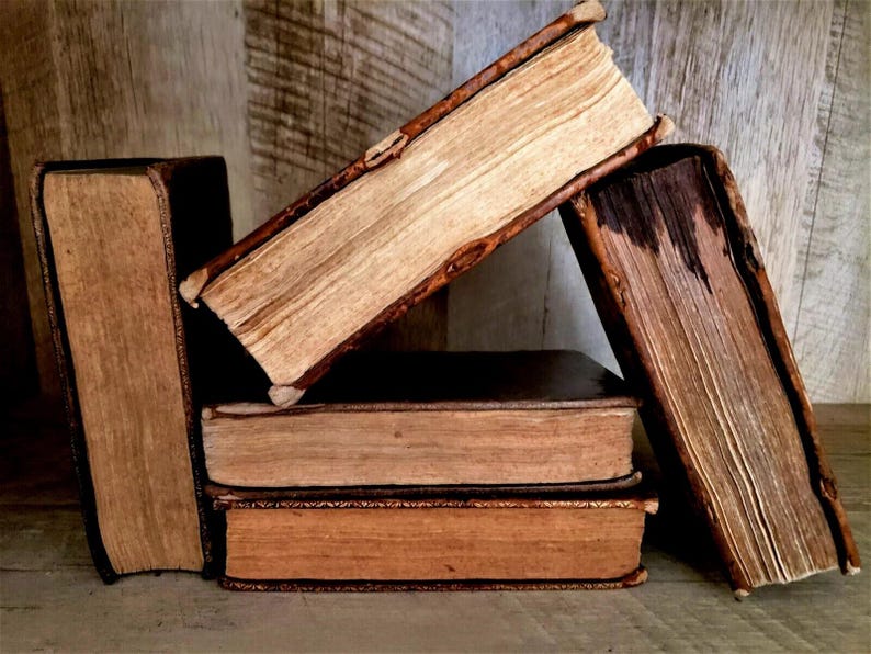 May include: A collection of antique books with aged, textured pages and worn, dark brown covers. The books are stacked and leaning against each other, creating a rustic and vintage aesthetic. The pages are a light tan color.