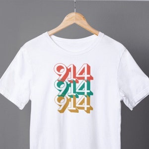 Puede incluir: Camiseta blanca con un diseño gráfico del número "914" repetido tres veces en diferentes colores: rojo, verde y dorado.