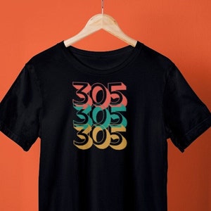 Puede incluir: Camiseta negra con un diseño gráfico del número 305 repetido tres veces en un diseño apilado y en capas. Cada número es de un color diferente: rojo, verde y amarillo.