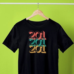 以下が含まれることがあります： 黒のTシャツに、2011年のグラフィックデザインが、ピンク、ターコイズ、イエローの3色で重ねてプリントされています。