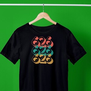 May include: Black t-shirt featuring a graphic design of the number 626 repeated three times in a stacked, offset pattern. The numbers are in different colours: red, teal, and yellow.