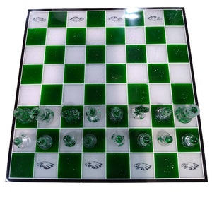 May include: A green and white chessboard with glass chess pieces. The chessboard has a green background with white squares and a black border. The chess pieces are made of clear glass with green accents. The chessboard has a design of an eagle on each side.