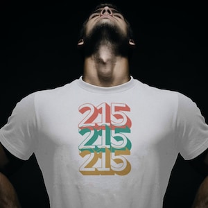 May include: A white t-shirt with a graphic design of the number 215 repeated three times in a stacked, layered, and slightly offset design. The numbers are in red, green, and yellow.