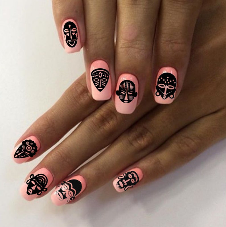 May include: Close-up of a hand with pink nail polish and black tribal mask nail art decals. The masks are different designs and are placed on each finger.