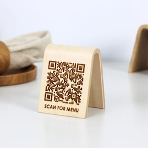 Puede incluir: Señales de mesa de madera con un código QR y el texto "SCAN FOR MENU". Las señales son de madera clara con una parte superior curvada y una base rectangular. Una señal está en posición vertical y otra está doblada. También se ven un servilletero de madera y una casa de madera.