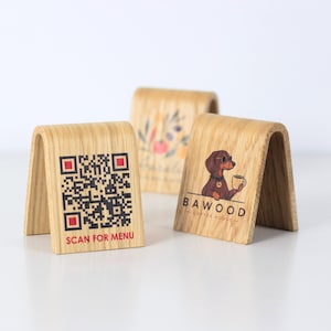 Puede incluir: Tres letreros de mesa de madera arqueada con acabado de veta clara. Un letrero presenta un código QR negro con detalles en rojo y el texto SCAN FOR MENU. Otro muestra una ilustración de un perro marrón con el texto BAWOOD COFFEE HOUSE.