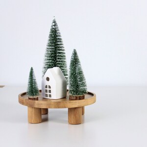 May include: A white ceramic house figurine sits on a wooden tiered stand with three small green artificial Christmas trees.