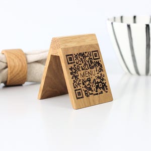 May include: A triangular wooden stand featuring a laser-engraved QR code and the word MENU on its front face. The light brown wood grain is visible across the structure, which is positioned next to a beige napkin ring and a white bowl with black stripes.