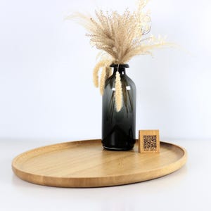 A minimalist wooden oval serving tray with a raised edge, featuring an amber glass bottle containing dried pampas grass and a small wooden block with a QR code placed on its surface. The tray is photographed against a plain white background.