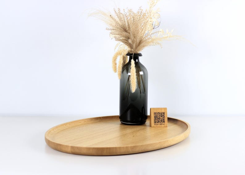 A minimalist wooden oval serving tray with a raised edge, featuring an amber glass bottle containing dried pampas grass and a small wooden block with a QR code placed on its surface. The tray is photographed against a plain white background.