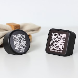 Puede incluir: Dos escáneres de códigos QR negros con el texto "SCAN FOR MENU". Uno es redondo, el otro es cuadrado. Una servilleta beige y un servilletero de madera están en el fondo.