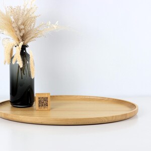 A minimalist wooden oval serving tray with a raised edge, featuring an amber glass bottle containing dried pampas grass and a small wooden block with a QR code placed on its surface. The tray is photographed against a plain white background.