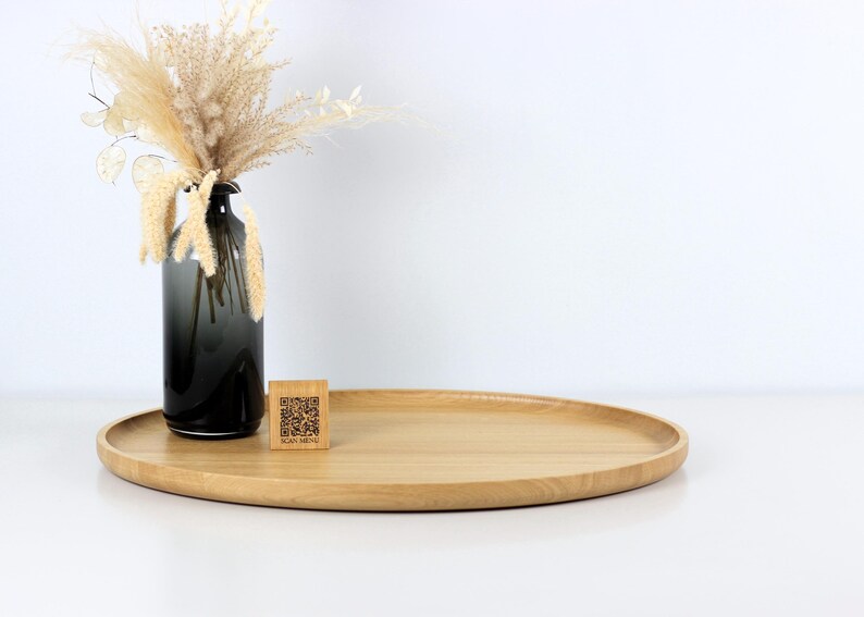 A minimalist wooden oval serving tray with a raised edge, featuring an amber glass bottle containing dried pampas grass and a small wooden block with a QR code placed on its surface. The tray is photographed against a plain white background.