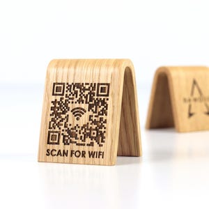 Può includere: Segno WiFi in legno con un codice QR e la scritta "SCAN FOR WIFI". Il cartello è in legno chiaro e ha una parte superiore curva. Un secondo cartello con un logo è sullo sfondo.