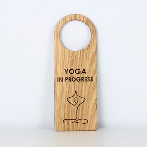 以下が含まれることがあります： 上部に丸い切り欠きのある木製のドアハンガー。「YOGA IN PROGRESS」の文字と、ヨガのポーズをとる人の線画が前面に刻まれています。木材は自然な木目を持っています。