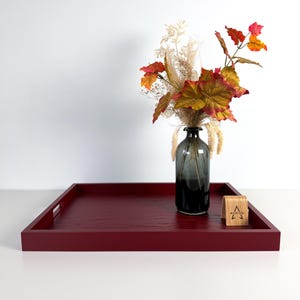 Pode incluir: Uma bandeja quadrada de madeira vermelho-escura com alças recortadas, apresentando um vaso de vidro cinza-escuro preenchido com folhas secas de cores outonais e gramíneas brancas, ao lado de uma pequena placa de madeira arqueada com o texto RA WOOD.