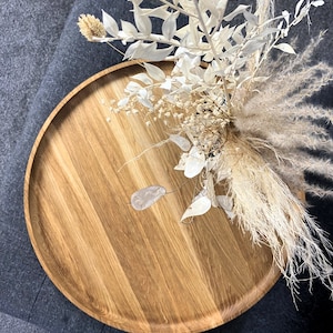 Round oak wooden tray styled on a grey ottoman with a black vase and dried floral arrangement, in a modern living room.