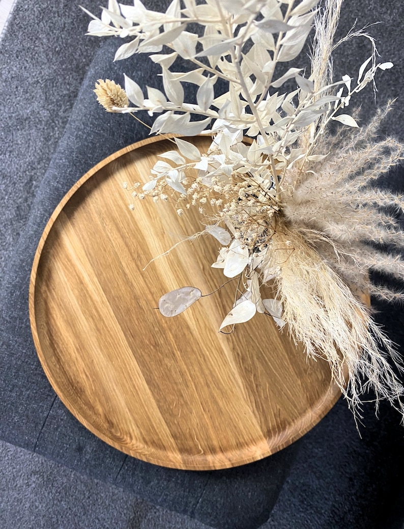 Round oak wooden tray styled on a grey ottoman with a black vase and dried floral arrangement, in a modern living room.