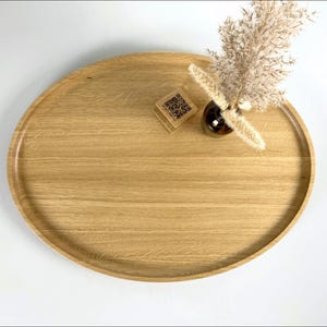 A minimalist wooden oval serving tray with a raised edge, featuring an amber glass bottle containing dried pampas grass and a small wooden block with a QR code placed on its surface. The tray is photographed against a plain white background.